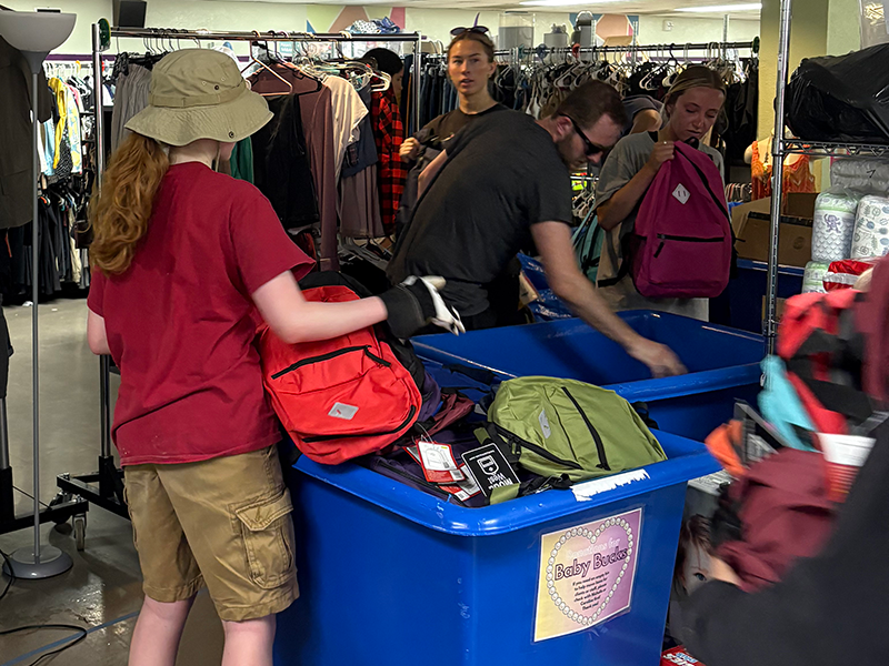 students organize backpacks into bins in front of racks of clothing