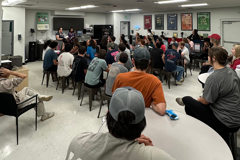 students seated at table listening to two speakers, standing