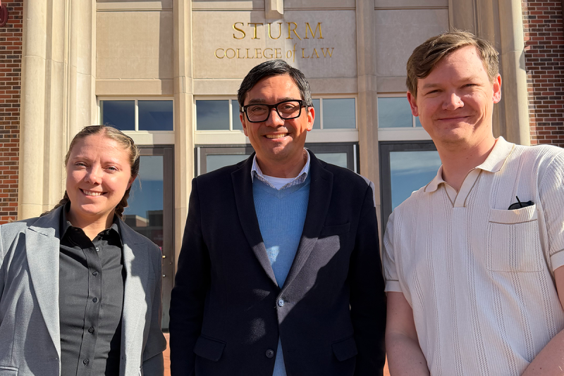 Three individuals standing in front of Sturm College of Law building
