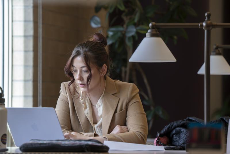 A female law student wearing a blazer works at a laptop computer in a law library.